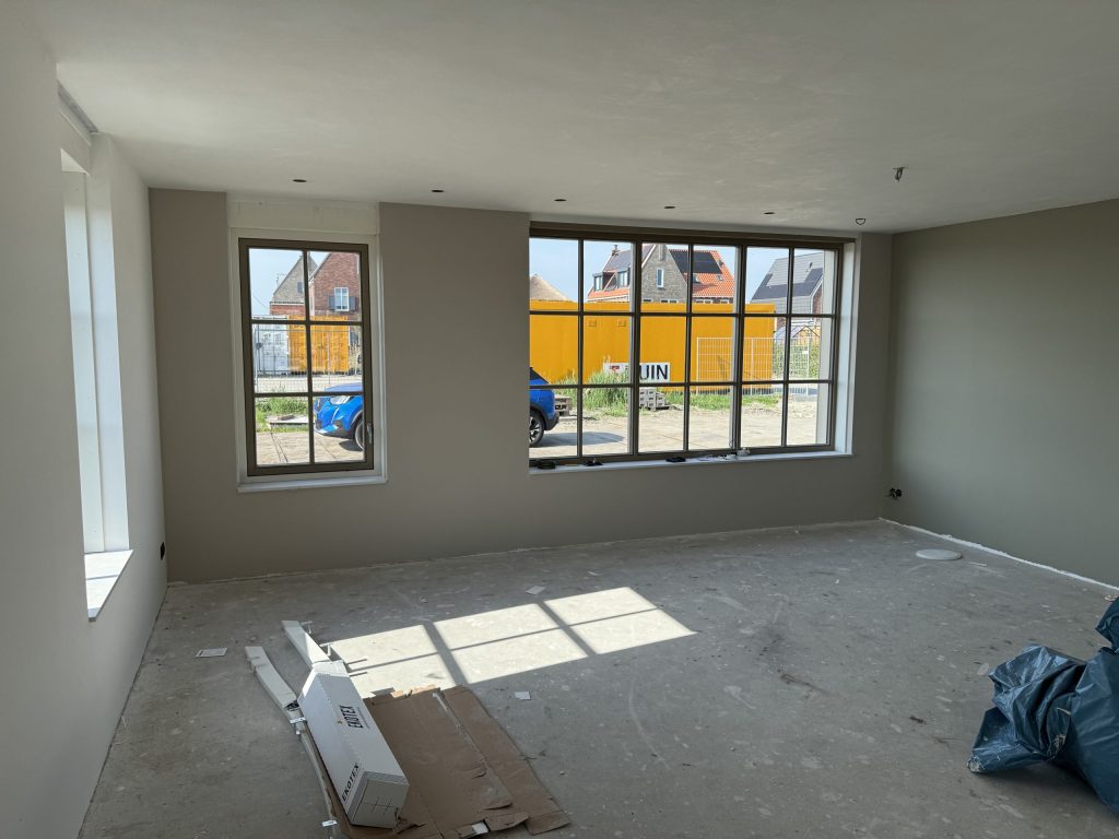 Lege nieuwbouwkamer met grijs gesausde wanden, grote raampartijen en een kale betonvloer