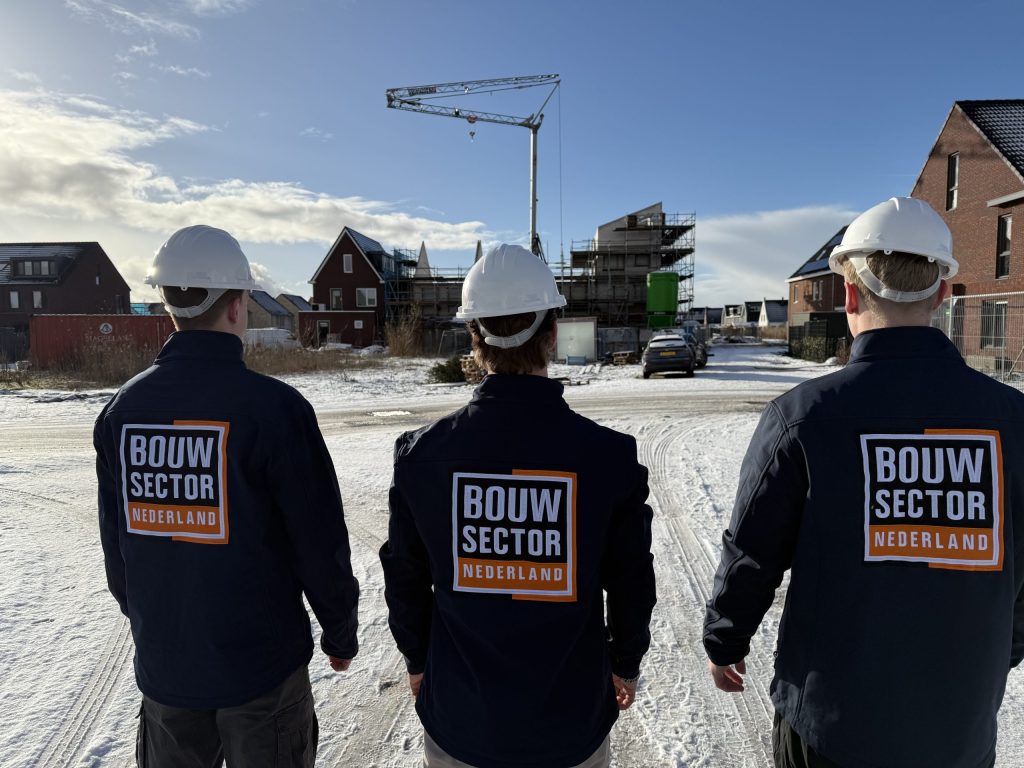 Drie vakmensen van Bouwsector Nederland met witte helmen en bedrijfsjassen lopen over een besneeuwde nieuwbouwlocatie met een bouwkraan